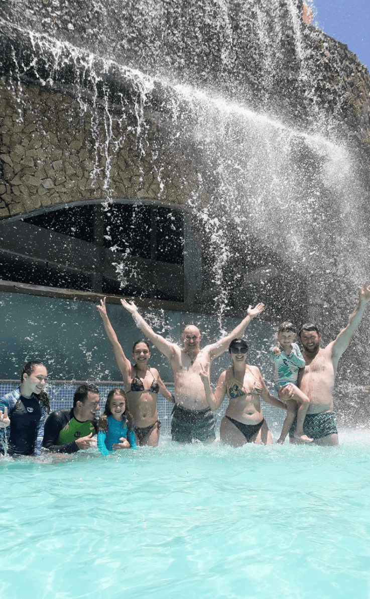 Uma família inteira se divertindo ao se banhar nas águas de uma cascata. Esposo, esposa, pai, mãe, filho, filha, sogro, sogra e sobrinhos.